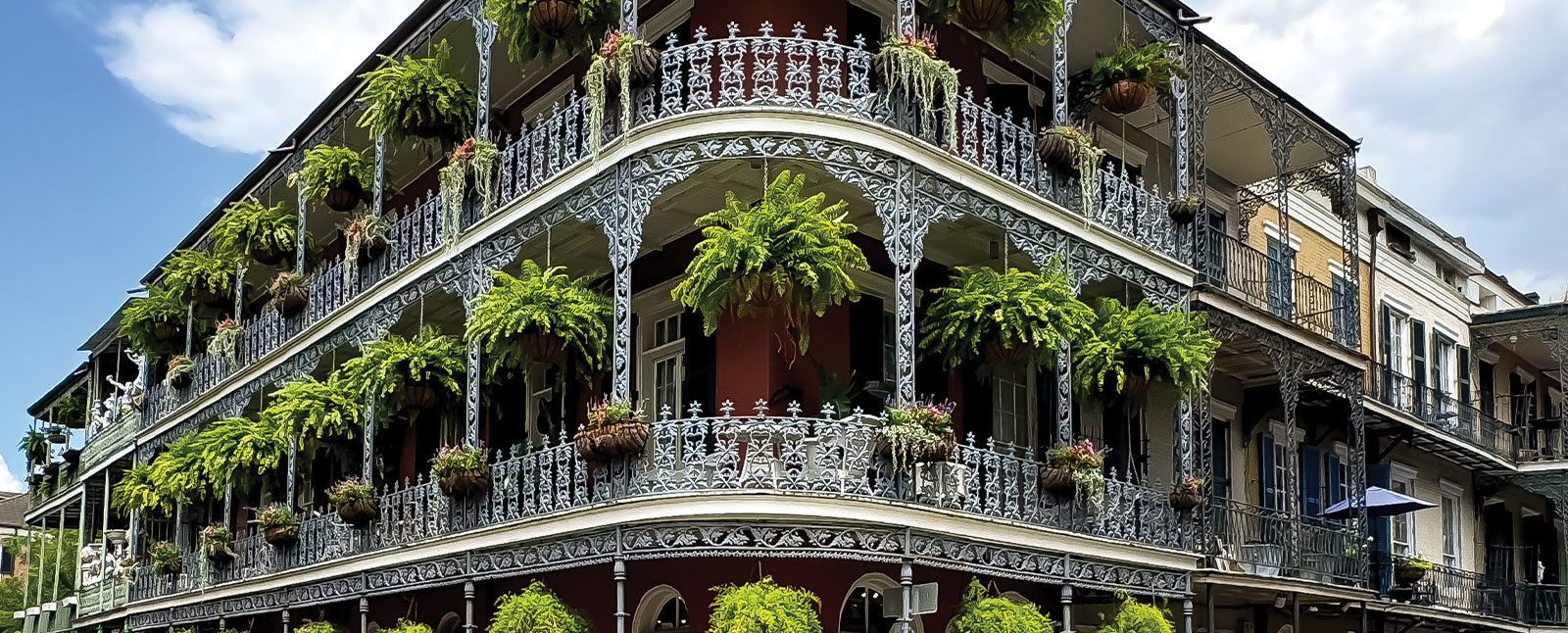 A corner building in New Orleans with ornate, white wrought-iron balconies decorated with lush green hanging ferns under a partly cloudy sky.