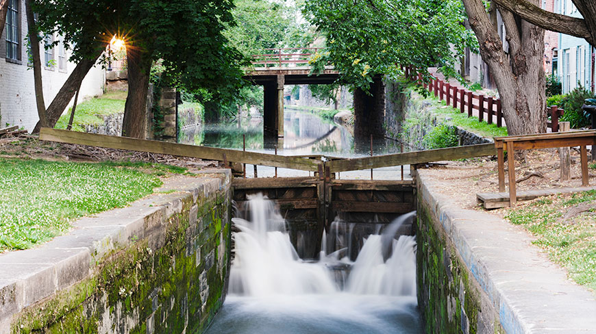 Water cascades through a historic lock on the C&O Canal in Georgetown, Washington, D.C., framed by lush trees and a distant bridge.