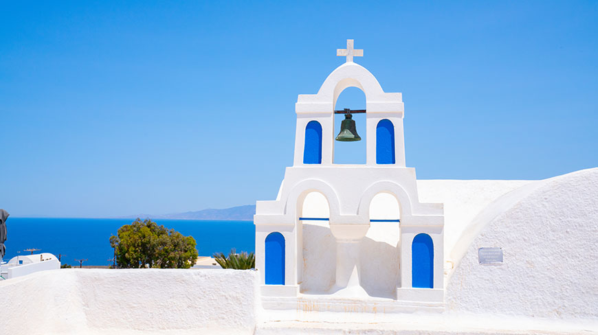 A whitewashed bell tower with bright blue details stands against the blue sea and sky in Santorini, Greece.