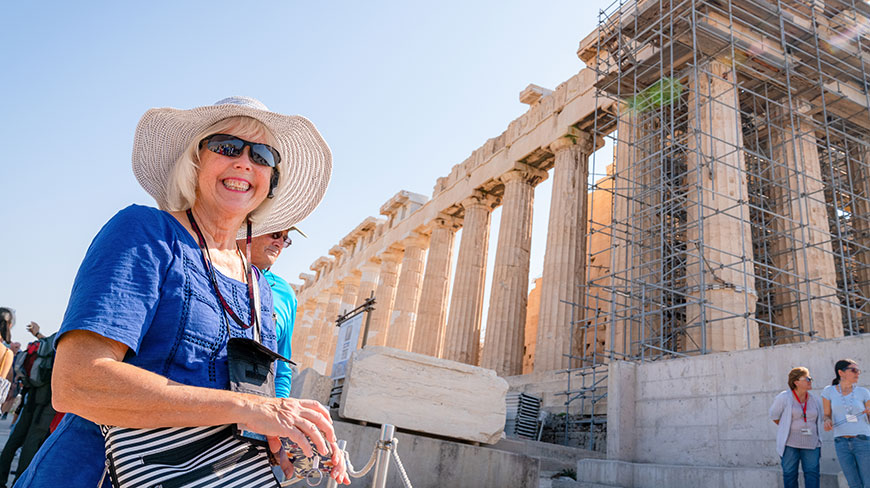 A smiling woman wearing a sun hat stands in front of the Parthenon on the Acropolis in Athens, Greece.
