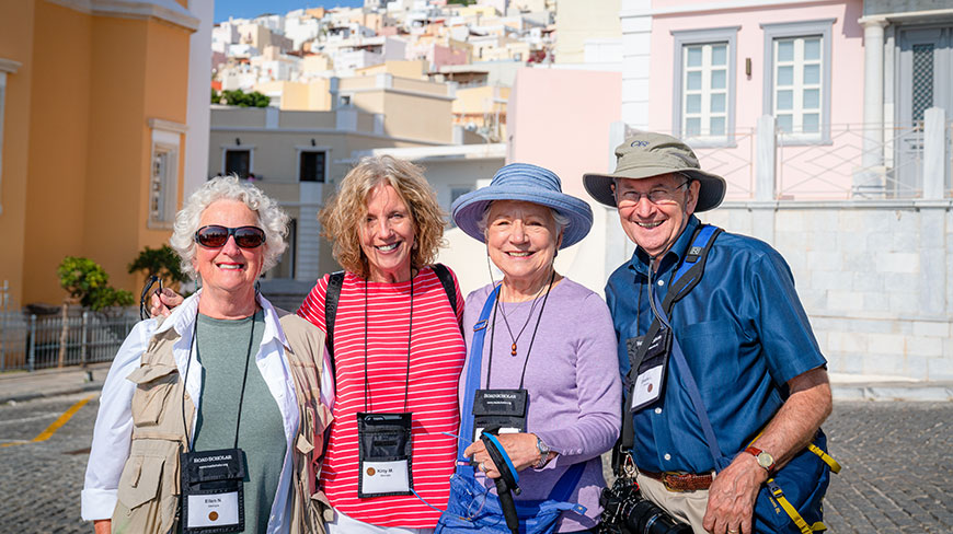 A group of four older adult travelers smile for a photo in a sunny Greek town with hillside architecture behind them.