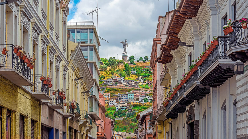A narrow street in Quito, Ecuador, is framed by colorful buildings, with the Virgen de El Panecillo statue visible on a distant hill.