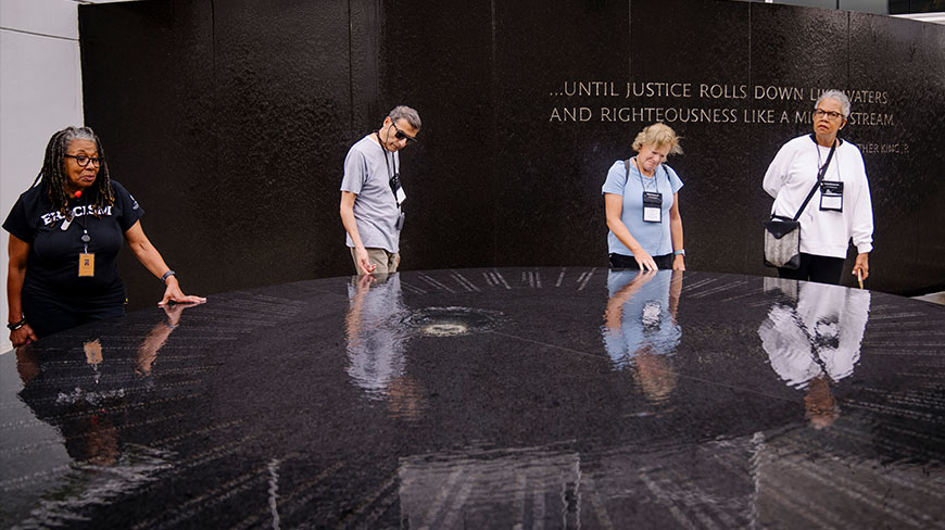 Four people gather around the wet, black stone of the Civil Rights Memorial in Montgomery, Alabama, with a memorial wall quoting Martin Luther King, Jr.