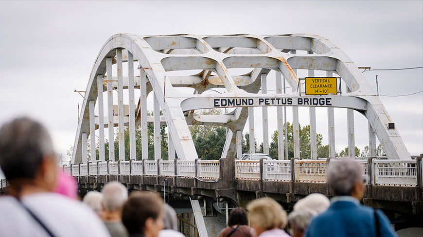 A crowd of people looks toward the large, white-arched Edmund Pettus Bridge in Selma, Alabama on an overcast day.