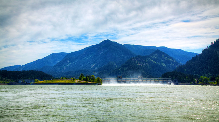 A wide view of the Bonneville Dam on the Columbia River in Oregon, with large, forested mountains rising in the background under a cloudy sky.