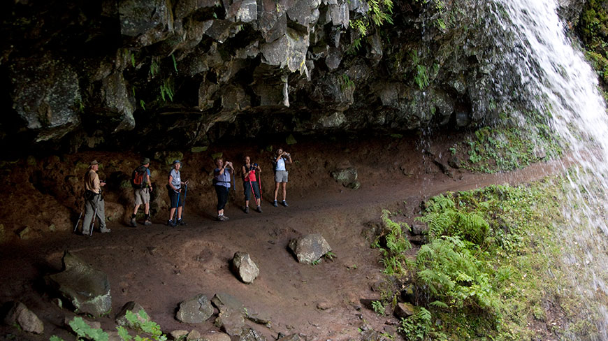 A group of hikers stand on a trail behind a waterfall in a rocky alcove in the Columbia River Gorge, Oregon.
