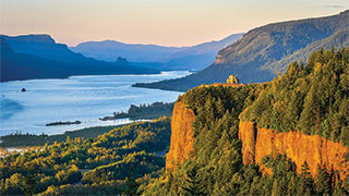 The sun sets over the scenic Columbia River Gorge in Oregon, casting a warm glow on the water and surrounding cliffs.