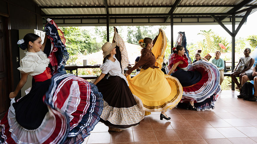 Women in colorful, traditional dresses perform a swirling folk dance for a small audience on a covered patio in Costa Rica.