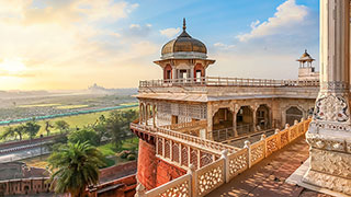 A beautiful stone balcony at the Agra Fort in India overlooks a hazy landscape with the Taj Mahal visible in the distance at sunrise.