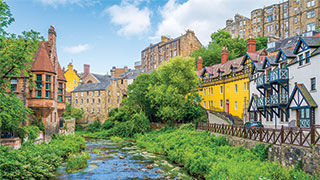 Colorful historic buildings line a lush, green riverbank in Dean Village, Edinburgh, Scotland, under a partly cloudy sky.