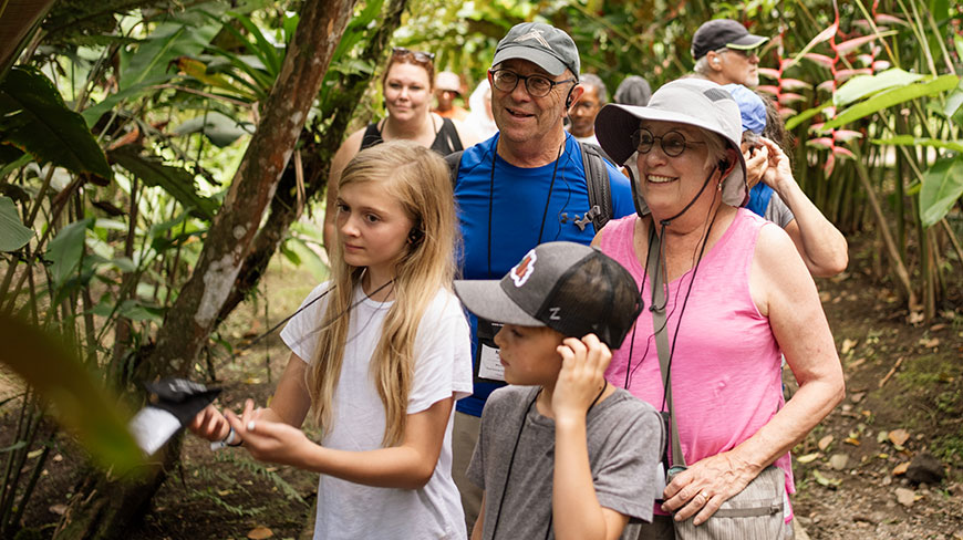 A family with grandparents and two children wear earpieces while on a guided nature walk through a lush forest in Costa Rica.