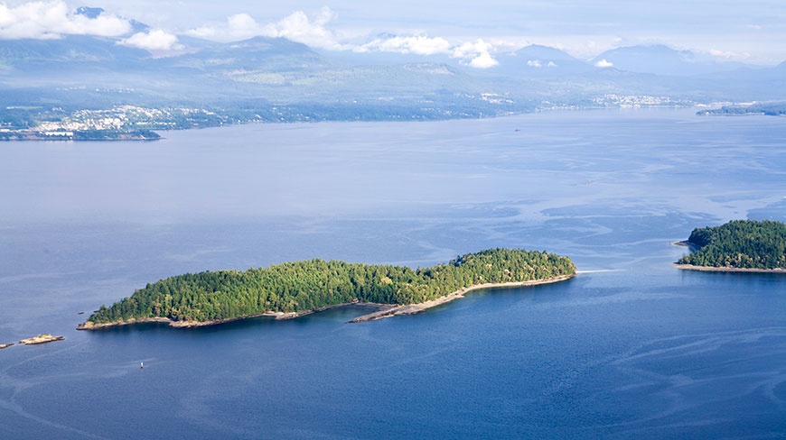 An aerial view of tree-covered islands dotting the blue waters of Alaska, with a mountainous coastline in the hazy distance.