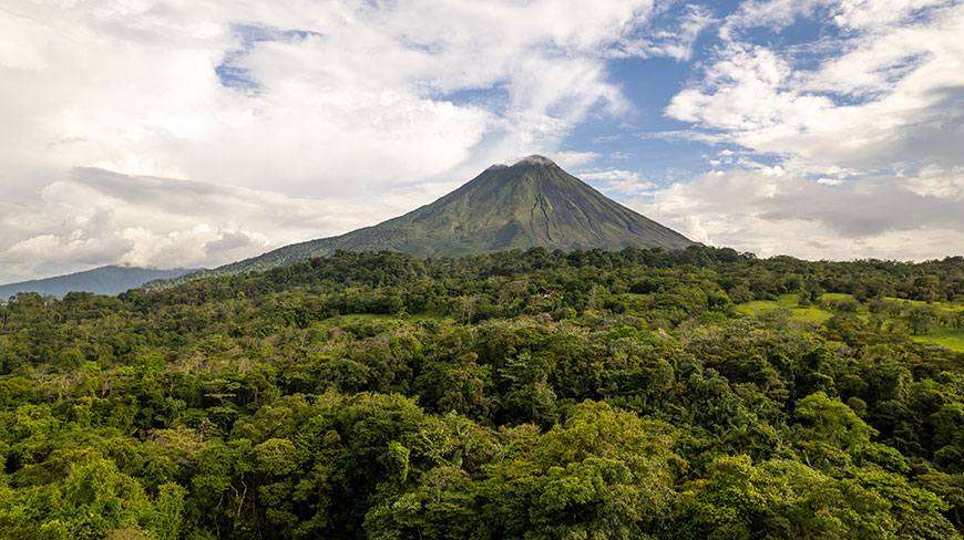 An aerial view of a volcano rising above a lush, green forest under a partly cloudy sky in Costa Rica.