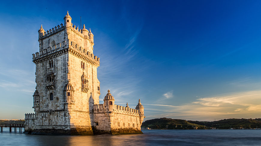 The historic Belém Tower in Lisbon, Portugal, sits on the water under a clear blue sky at sunset.