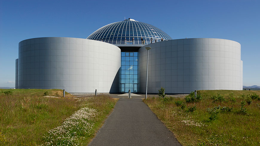 The Perlan Museum in Iceland, a domed building atop large cylindrical tanks, viewed from a path through a grassy field under a clear blue sky.