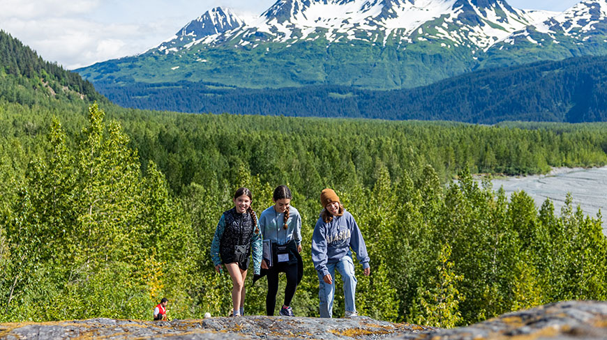 Three young people hike on a rocky trail in Alaska with a lush forest and snow-capped mountains in the background.