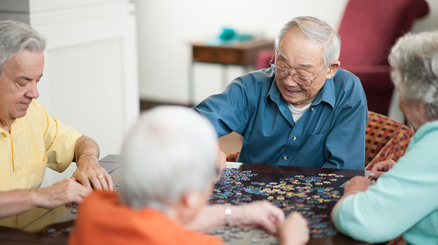 A group of smiling seniors works together on a jigsaw puzzle at a table in a community living room.