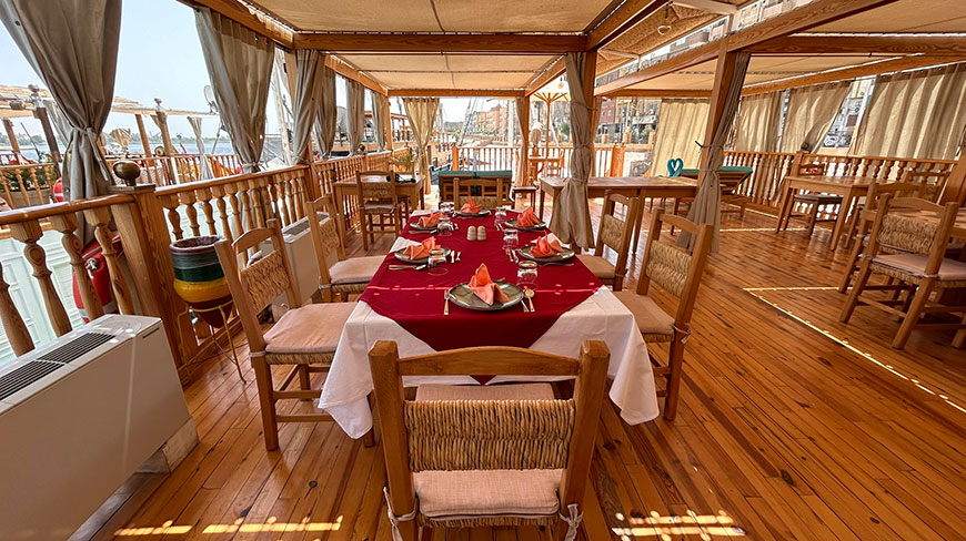 A dining table set with a red tablecloth on the outdoor wooden deck of a dahabeya boat sailing along the Nile River in Egypt.