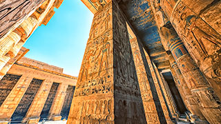 A low-angle view of the sunlit, hieroglyph-covered columns at Medinet Habu Temple in Luxor, Egypt.