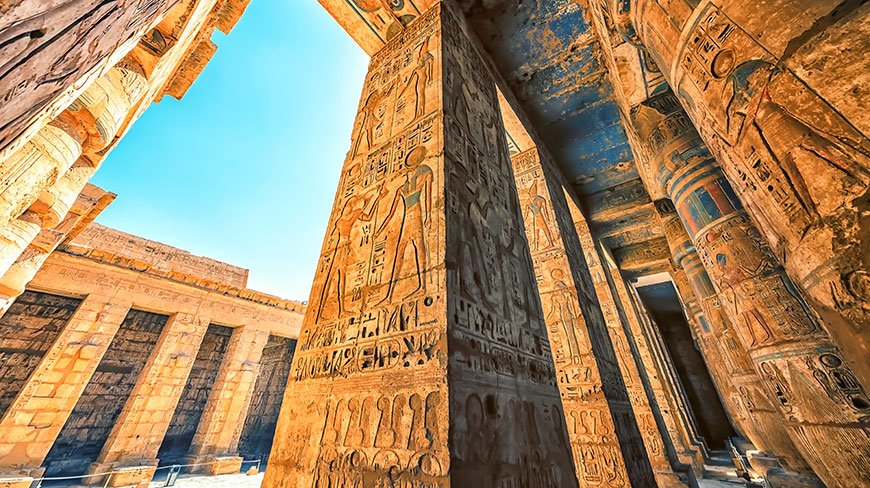 A low-angle shot looking up at the sunlit, hieroglyph-covered columns of the Habu Temple in Luxor, Egypt, against a clear blue sky.