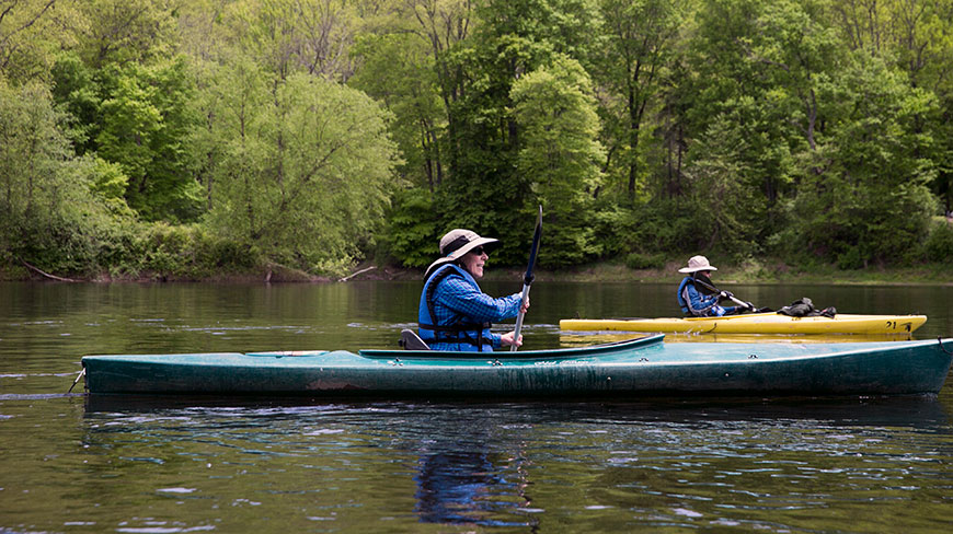 Two women paddle in kayaks on a calm river in Pennsylvania, with a dense green forest on the far shore.