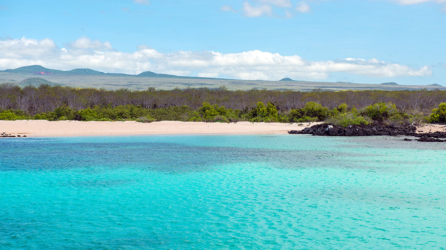 Turquoise water washes up on a sandy beach in the Galápagos Islands, with volcanic hills visible in the distance.