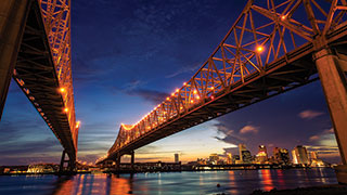 The Crescent City Connection bridge in New Orleans, Louisiana, illuminated at dusk as it spans the Mississippi River with the city skyline behind it.