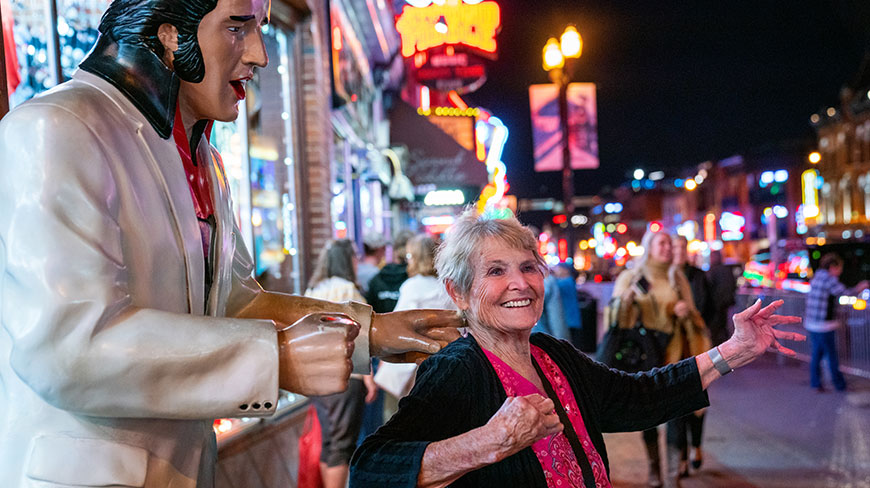 A smiling woman dances with a statue of Elvis Presley at night on a crowded, neon-lit Beale Street in Memphis.