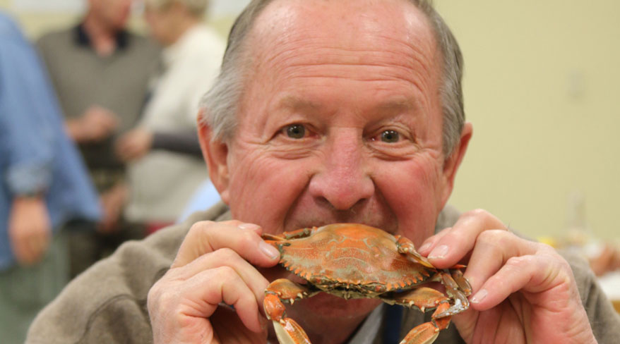 A smiling man holds a whole cooked crab to his mouth to take a bite during a sea-to-table event in Virginia.