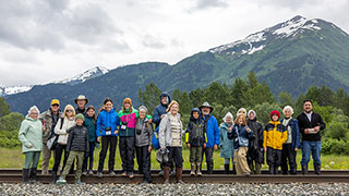 A group of travelers on a family adventure stand on railroad tracks with snow-capped mountains in the background in Alaska.