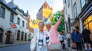 Two women wave happily on a cobblestone street in Québec, with the grand Château Frontenac visible in the background.