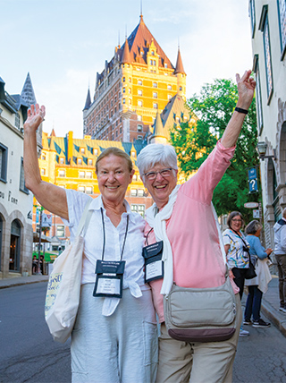 Two happy women wave on a street in Québec City, with the historic Château Frontenac visible in the background.