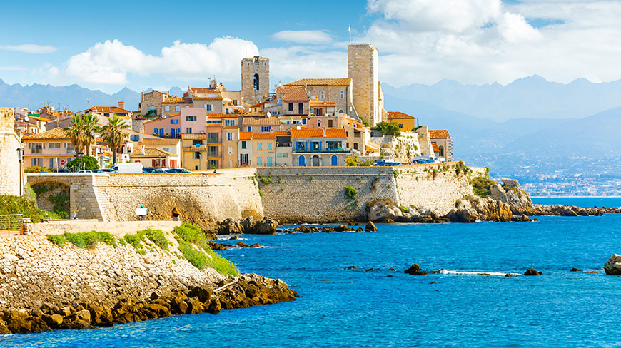 The historic center of Antibes, France, is seen from the bright blue Mediterranean Sea with mountains in the background.