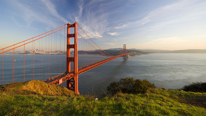 The Golden Gate Bridge in San Francisco is viewed from a grassy hill at sunset, with the city skyline visible across the bay.
