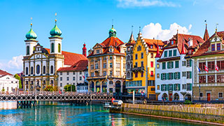 Colorful historic buildings and a church with green domes line a river in Lucerne, Switzerland on a sunny day.