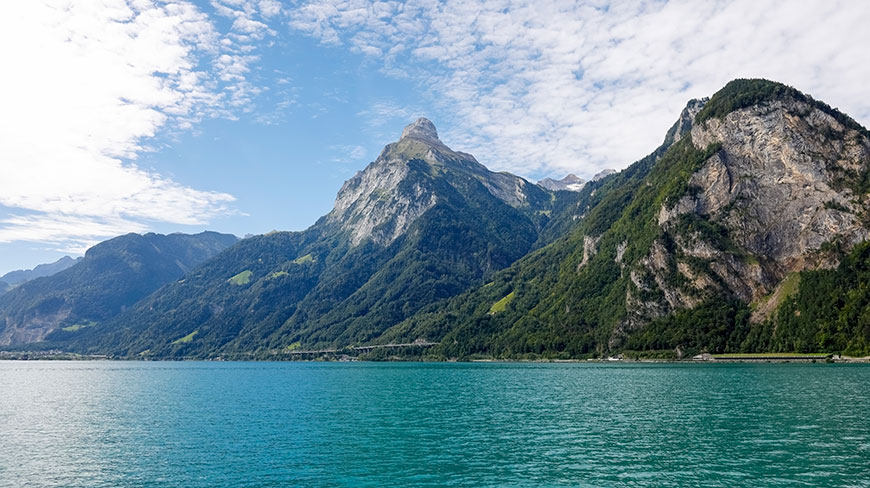 The turquoise water of Lake Lucerne in Switzerland is surrounded by steep, forest-covered mountains under a blue sky with white clouds.