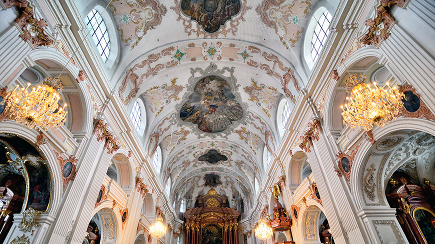 The ornate, painted ceiling of the Jesuit Catholic Church in Switzerland, with large chandeliers hanging from the vaulted arches.