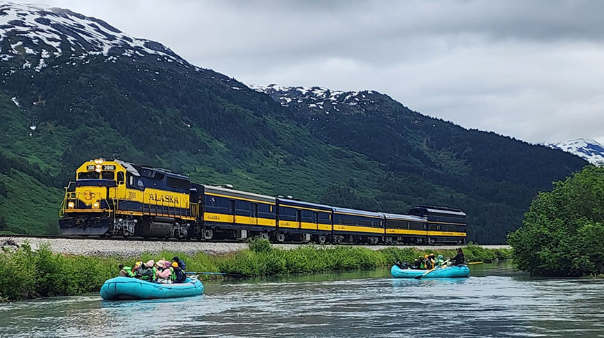 People in rafts float down the Placer River in Alaska as a blue and yellow train passes by with mountains in the background.