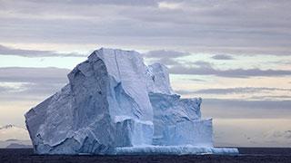 A large, jagged iceberg with steep cliffs of blue and white ice floats on the dark ocean water under a cloudy sky.