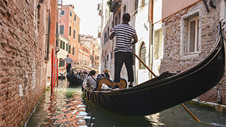 A gondolier steers a gondola with passengers down a narrow canal lined with old brick buildings in Venice, Italy.