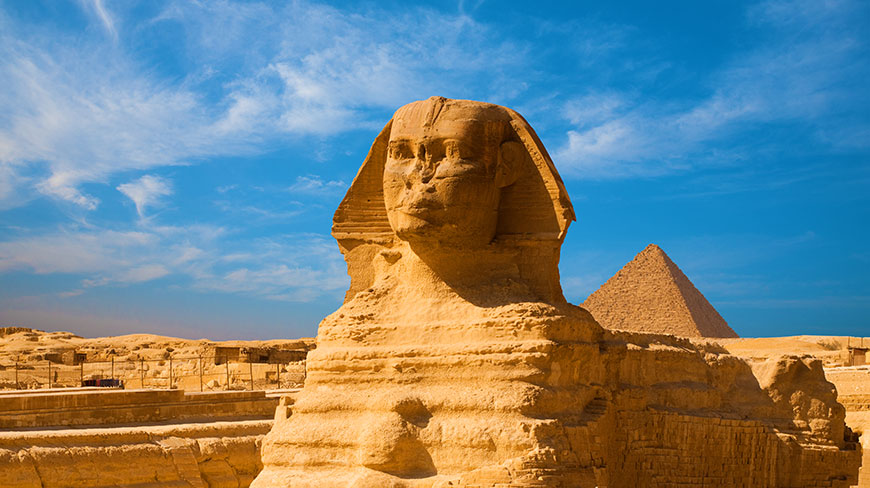 The Great Sphinx of Giza sits under a bright blue sky, with a large Egyptian pyramid visible in the background.
