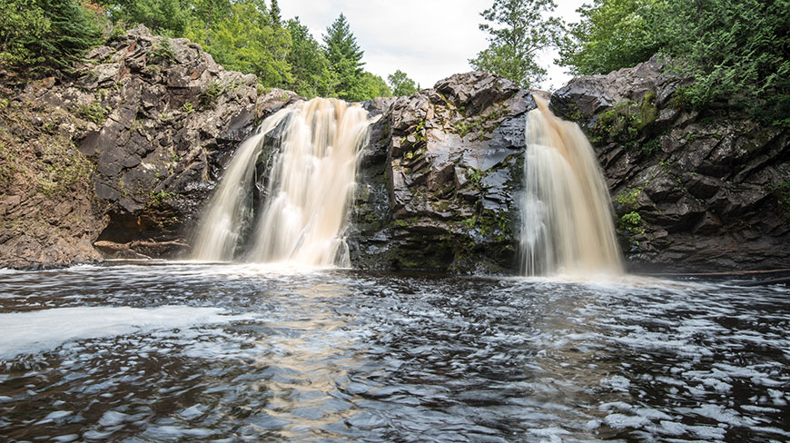 Two waterfalls cascade over dark, rocky cliffs into a churning pool of water at Pattison State Park in Wisconsin.