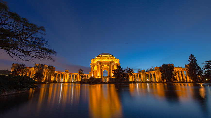 The Palace of Fine Arts in San Francisco illuminated at dusk, its golden lights reflecting in the calm water of the lagoon.