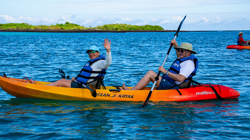 Two people in a tandem kayak paddle on the ocean in the Galápagos Islands, as one person waves at the camera.