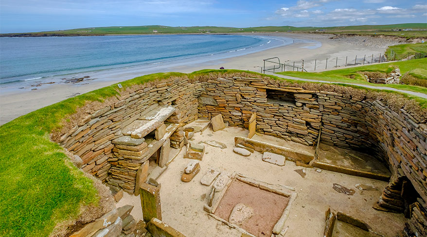 The ancient stone settlement of Skara Brae, with grass-topped walls, overlooks a wide sandy beach and the ocean in Orkney, Scotland.