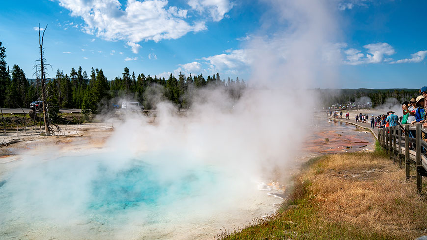 Tourists on a boardwalk view a large, steaming turquoise geothermal pool in Yellowstone National Park, Wyoming under a partly cloudy blue sky.