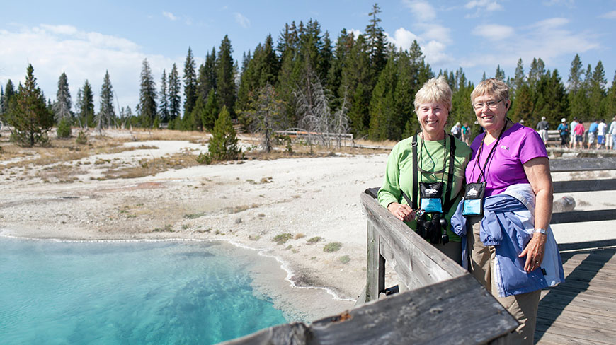 Two women smile on a boardwalk next to a bright blue hot spring in Yellowstone National Park in Montana/Wyoming.