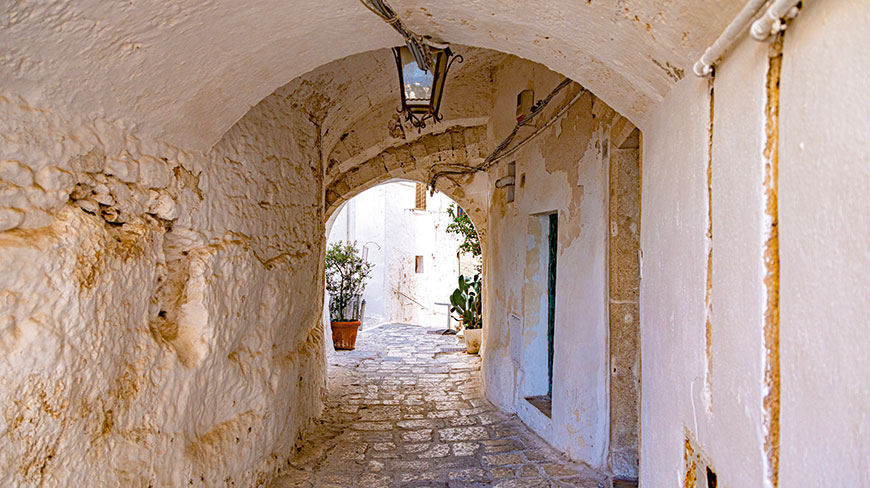 A narrow cobblestone alley with whitewashed, arched stone walls leads to a sunlit area with potted plants.