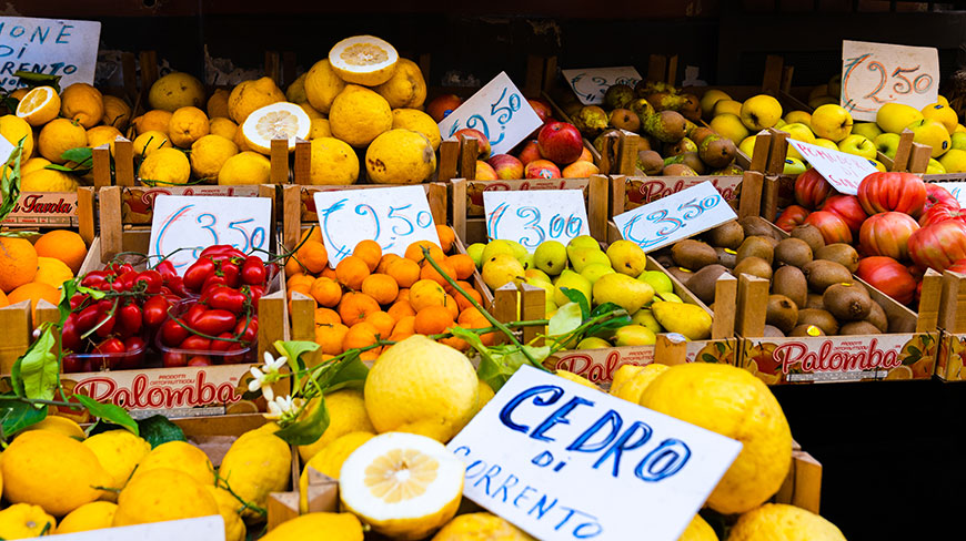 A colorful market stall displays a variety of fresh fruits like lemons, pears, and kiwis in wooden crates with handwritten price signs.