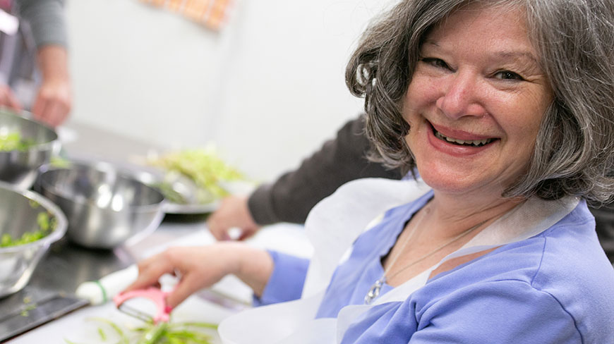 A smiling woman with gray hair wearing a blue shirt and apron peels vegetables during a cooking class.